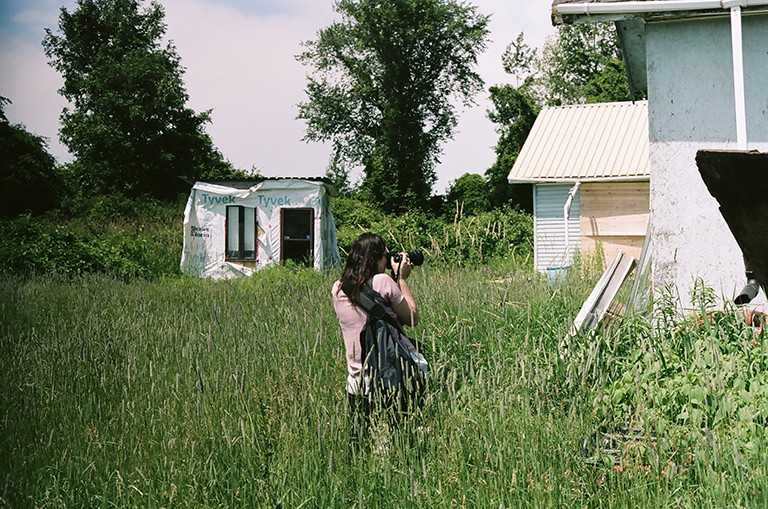 Une personne munie d'un appareil photo se tenant dans un champ d'herbes hautes et photographiant des bâtiments abandonnés.