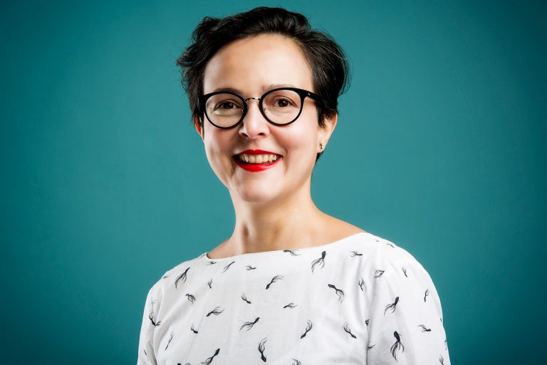 A light-skinned woman with dark short hair and glasses, wearing a white patterned shirt, red lipstick and smiling against a dark turquoise background.
