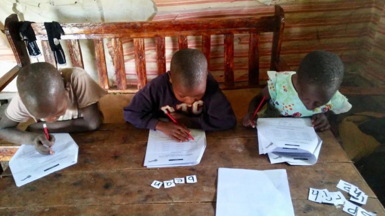 Three children from the Trans Mara region of Kenya sit at a desk doing their schoolwork.