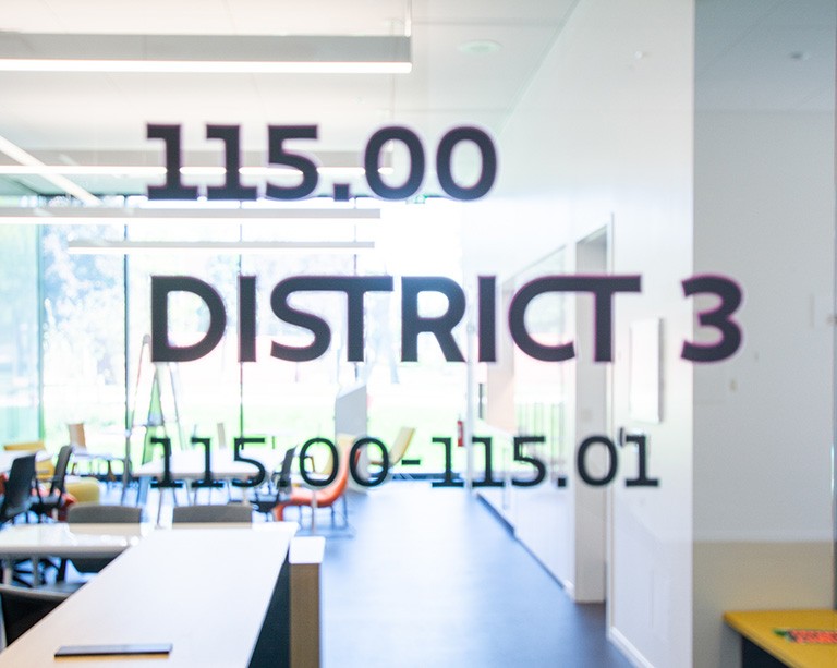Blurry view through a window with the words "155.00, District 3, 115.00-115.01" on the glass, into a classroom.