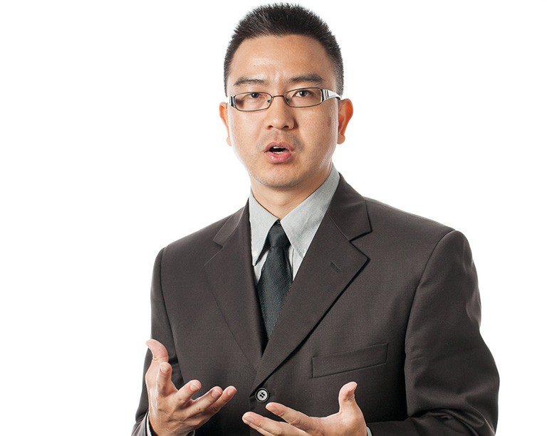 A young man with short dark hair, of asian heritage, wearing steel-rimmed glasses and a brown suit.