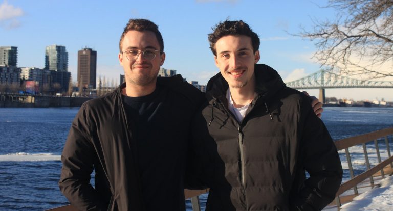 Two young men standing together, with a river and city in the background.