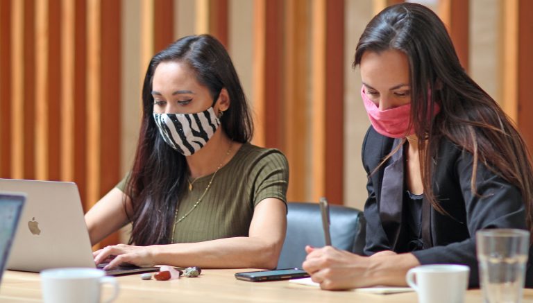 Two women with long dark hair, wearing masks and working at laptops.