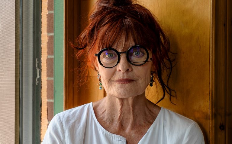 An older woman with a white shirt, long red hair piled on top of her head and round, black-rimmed glasses.