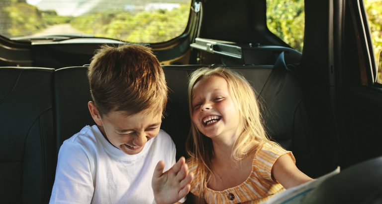 Pictured: Two young children, around 6 and 7 years old, that look like brother and sister. They're sitting in the back seat of a car and the young girl has said something that is making them laugh really hard.