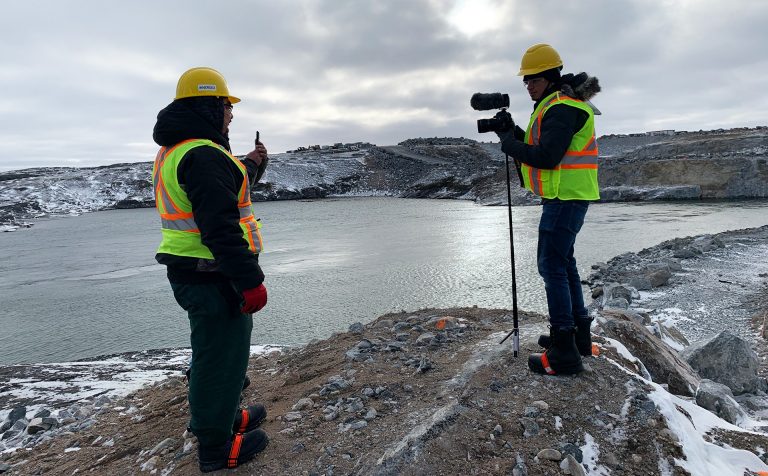 Two men in winter gear and hard hats standing on a rocky outcrop before a large body of water. One man has a camera on a tripod.