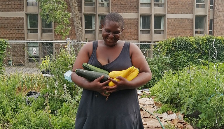 Smiling Black woman in a black, sleeveless dress, holding courgettes in a community garden.