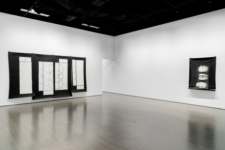 Installation view of and exhibition, with a series of drawings hung on black paper with white walls behind.