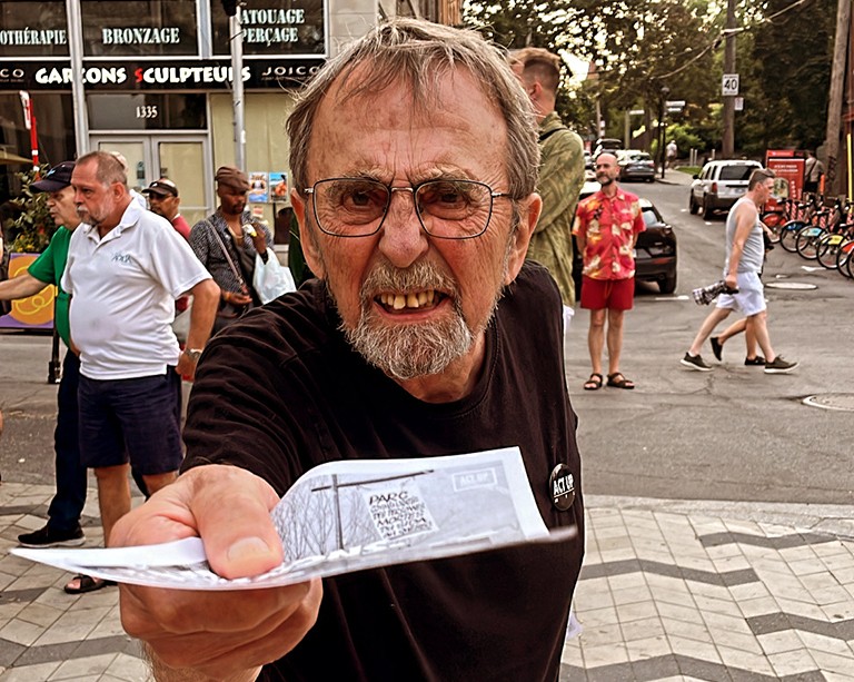 An angry looking middle-aged man in a black T-Shirt and with short, blonde hair and glasses holding out a paper flyer to the person taking the photograph.