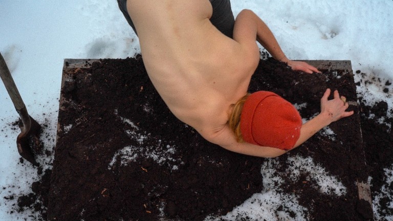 Shirtless person with red toque and back to camera grabbing at earth in gardening plot. There is snow around the plot.