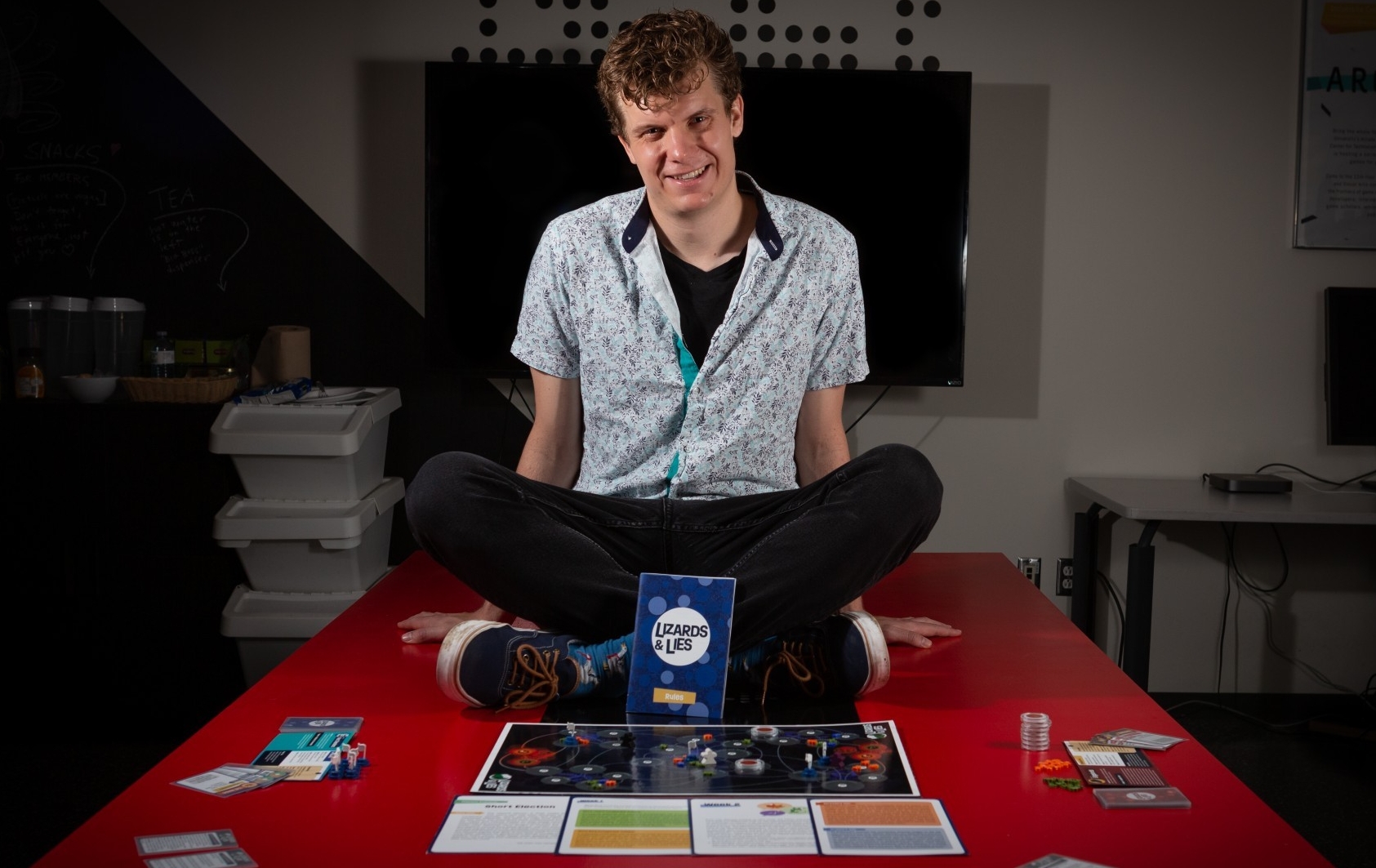Young man with short, curly blonde hair, wearing a shirt sleeved shirt and black jeans and sitting on a table with a board game called "Lizards and Lies" spread out in front of him.