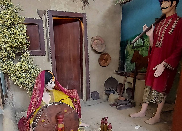 Diorama at a museum depicting a figure of a woman in traditional dress seated on the floor and a figure of a standing mustached man in traditional dress.