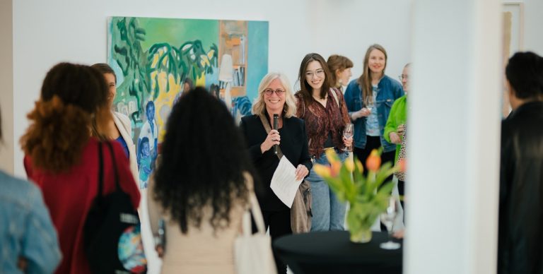 A group of people listening to a woman with blonde hair and glasses at a vernissage for an art show.