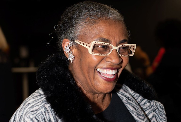 Smiling woman with pulled back, black and grey hair, with beige and tan-rimmed glasses and wearing a black and white jacket with black fur trim.