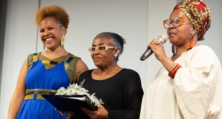 Three smiling women standing on a stage facing an audience, the woman in the middle holding an award and the woman on the far right speaking into a microphone.