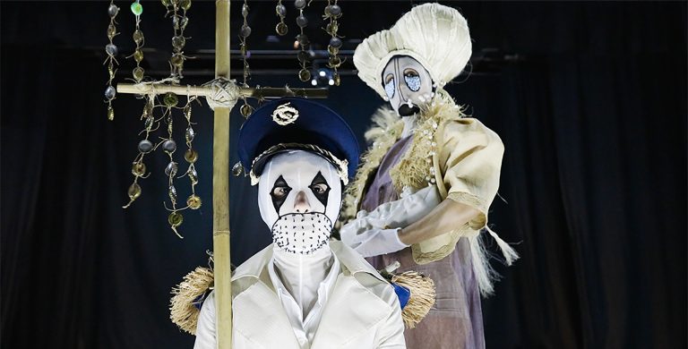 Two figures wearing white and beige costumes and masks with eyes cut out.