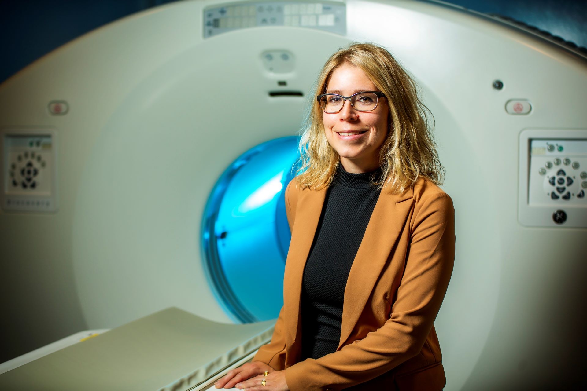 Maryse Fortin wears a brown jacket over a black shirt and sits in front of an MRI machine