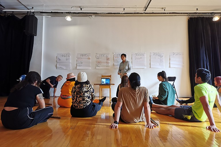 A group of people sitting on the floor of a dance studio and listening to a presentation.