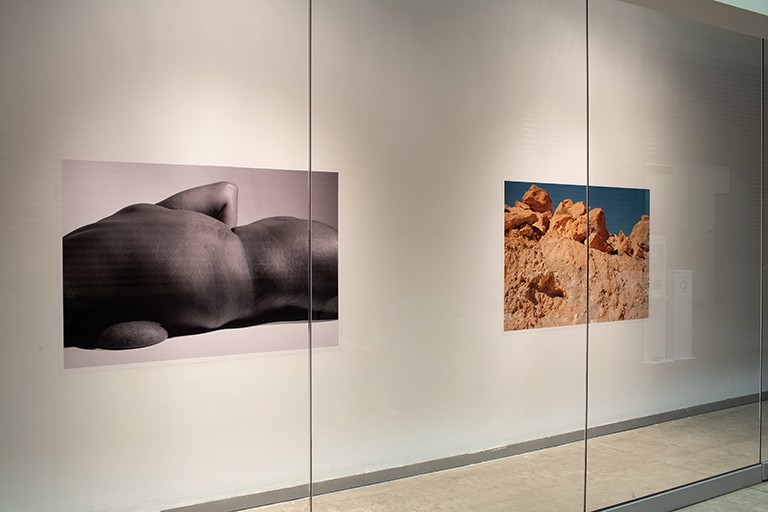 View of a gallery wall with two different artworks, both photos, one of a naked torso and one of red mountains.