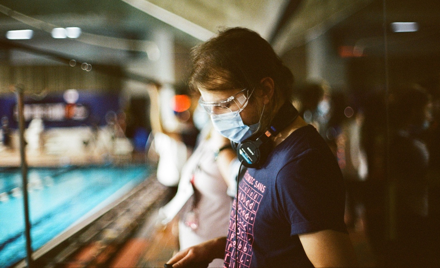 Un homme porte des écouteurs, un t-shirt et un masque chirurgical sur le plateau de tournage, à côté d'une piscine.