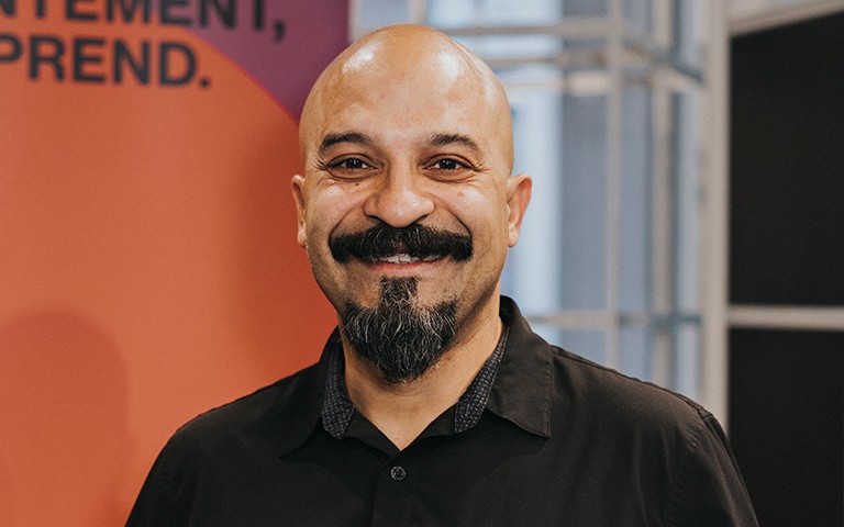 Homme souriant au crâne rasé, à la barbe sombre et portant une chemise noire.