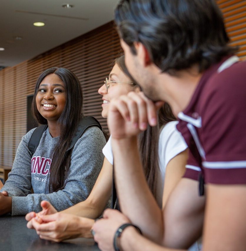 Three students in a campus setting converse among themselves. Three students in a campus setting converse among themselves.