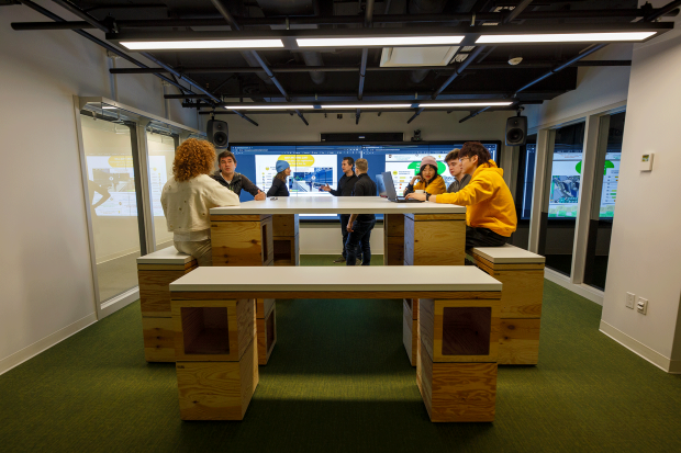 Students sit at a table with a laptop and a screen behind them