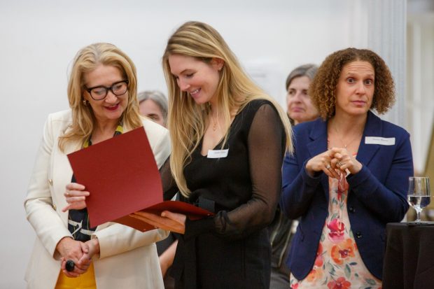 A woman shows a red folder to another woman while a group watches in the background at an indoor event.