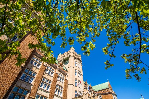 A historic brick building with a tower is seen from below, framed by green leaves under a bright blue sky.