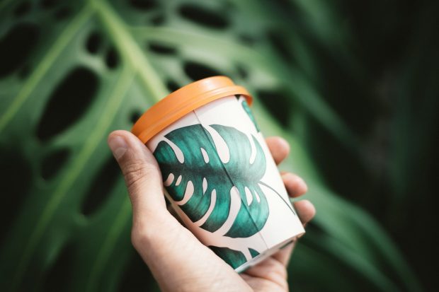 Hand holding a cup with a green leaf pattern and an orange lid, with a large leaf in the background.