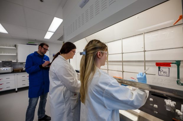 Three scientists wearing lab coats work at a laboratory fume hood, conducting experiments with lab equipment on a counter.