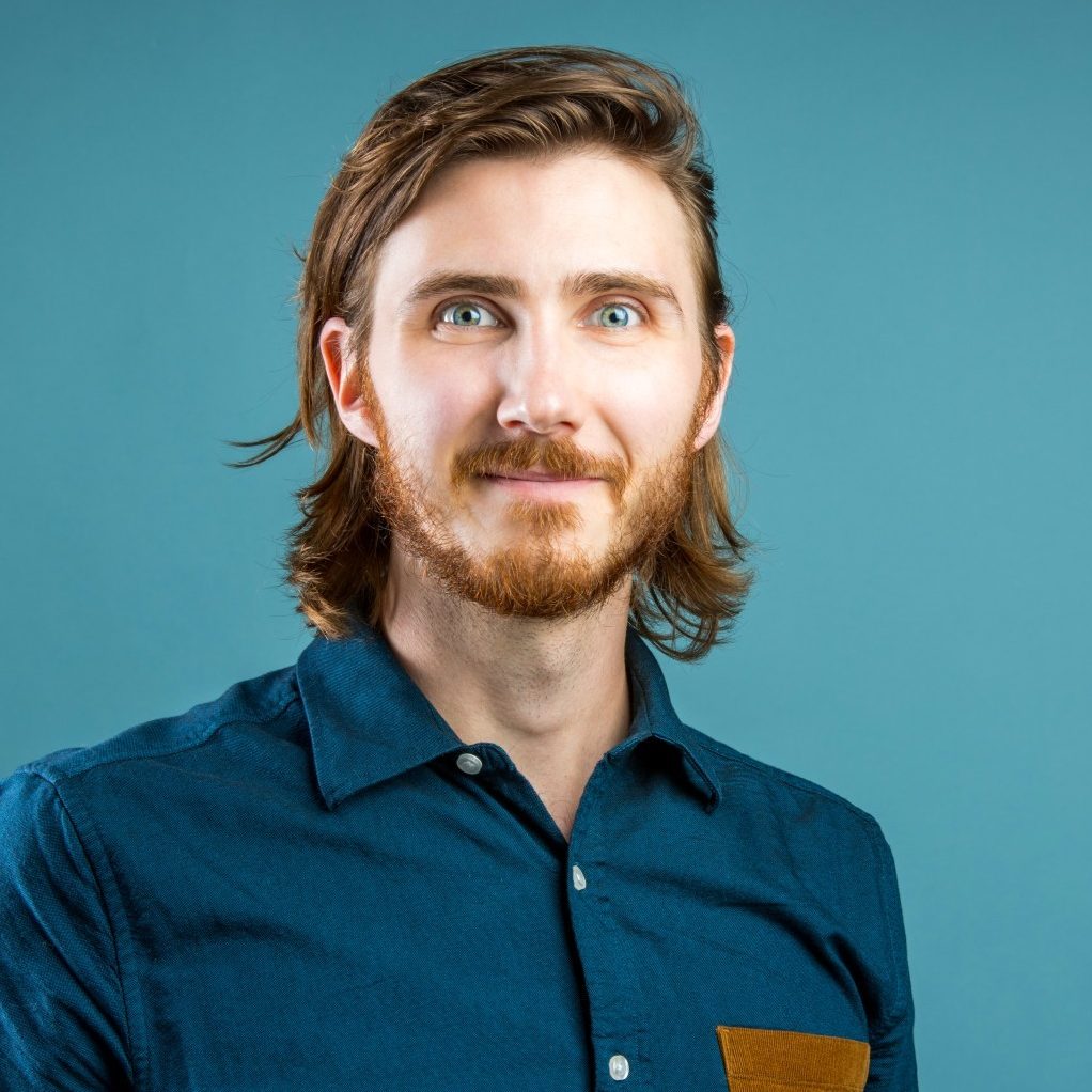 A man with medium-length hair and a beard is wearing a teal shirt with a brown pocket, against a blue background.