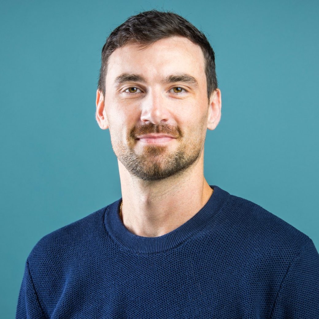 A man with short dark hair and a beard is wearing a navy sweater, standing against a teal background.