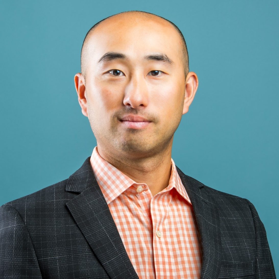 A man in a checked shirt and dark blazer poses in front of a blue background.