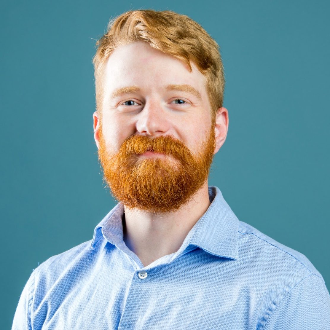 A person with red hair and a beard is wearing a light blue button-up shirt against a blue background.