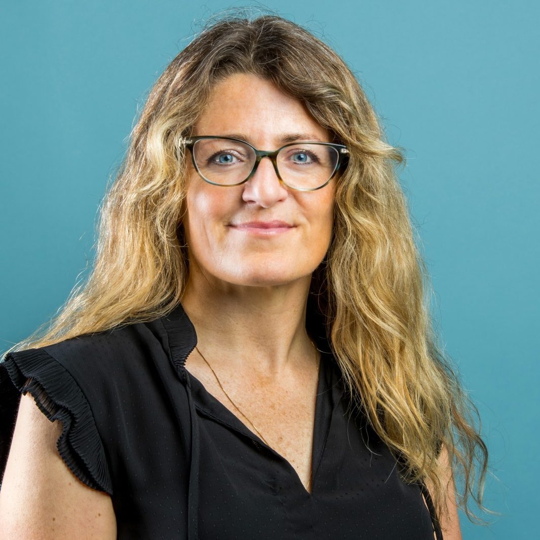 A person with long, wavy hair and glasses, wearing a black shirt, stands against a teal background.