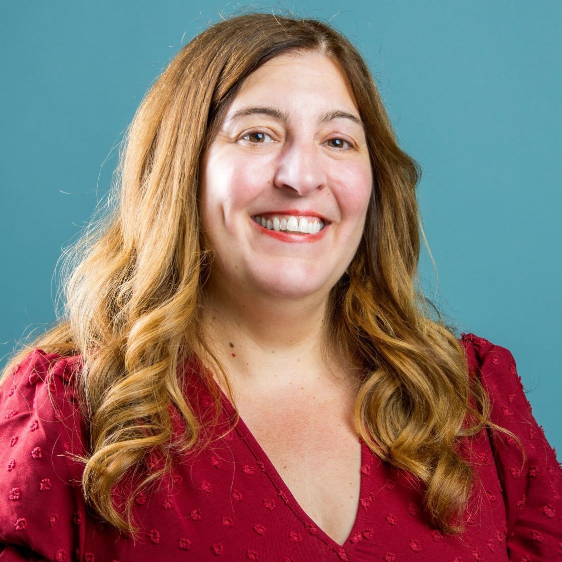Woman with long hair wearing a red blouse, smiling against a teal background.