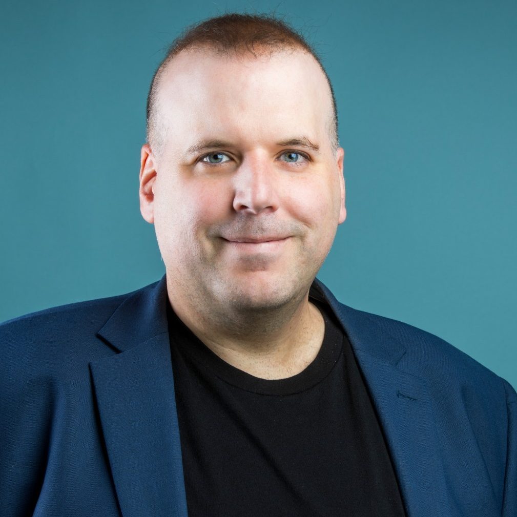 A person in a blue blazer and black shirt smiles against a teal background.