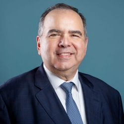 A person in a suit and tie smiles against a blue background.