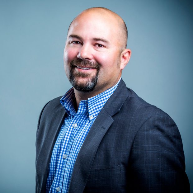 D'Arcy Ryan A man with a beard and mustache, wearing a blue checkered shirt and dark suit jacket, smiles against a plain gray background.