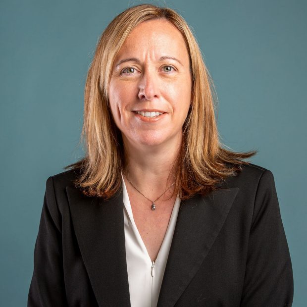 A person with shoulder-length hair is smiling, wearing a black blazer over a white top, against a plain teal background.