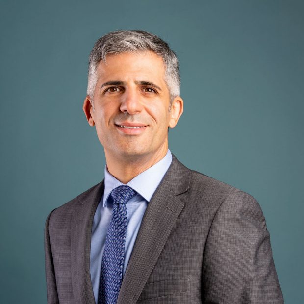 A man in a grey suit and blue tie stands against a teal background, smiling at the camera.