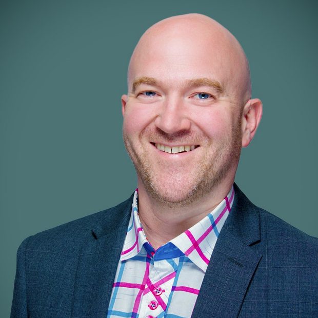 A man wearing a blue suit and a colourful checkered shirt smiles against a green background.