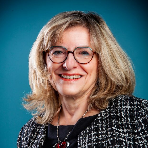 A smiling woman with glasses and shoulder-length blonde hair, wearing a black and white jacket, poses in front of a blue background.