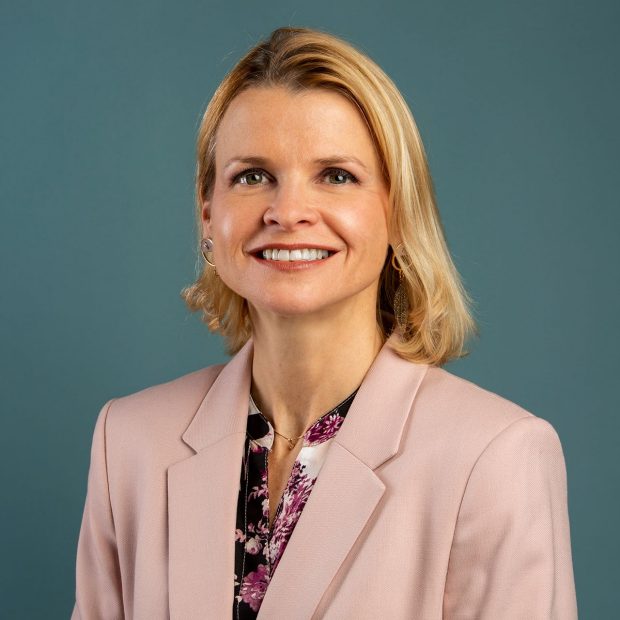 A person with shoulder-length blonde hair smiles, wearing a light pink blazer over a floral top, against a plain teal background.