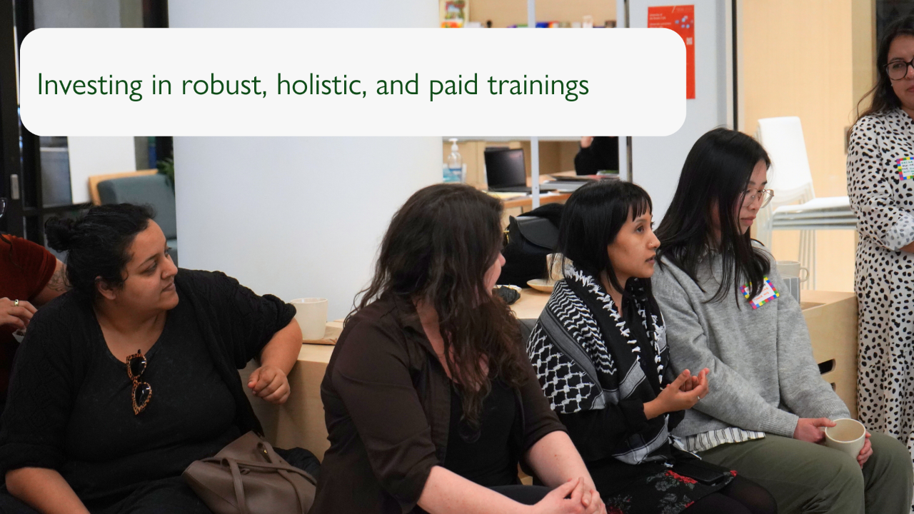 Slide with a photograph of an audience gathered at an event, with one person speaking. The slide reads "Investing in robust, holistic, and paid trainings".