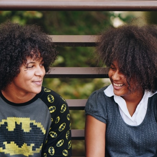 Two Black youth sit side-by-side looking at each other with shy smiles