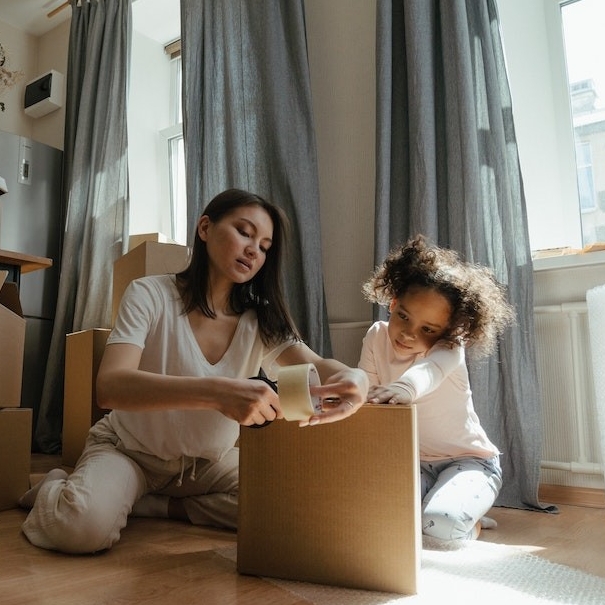A mother and child sit on the floor taping a box closed as they prepare to move