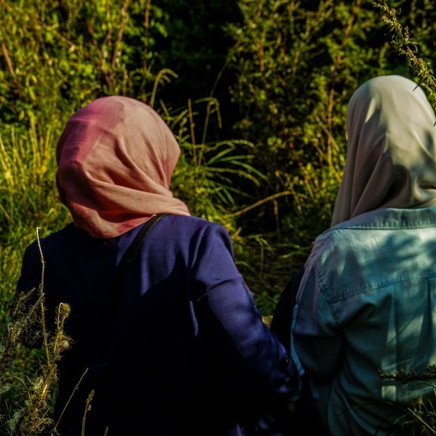 A view from behind of two women wearing hijabs walking arm and arm in a forest.
