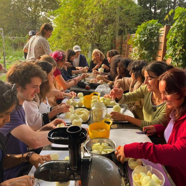 A photograph of a group sitting at a large table outside peeling apples
